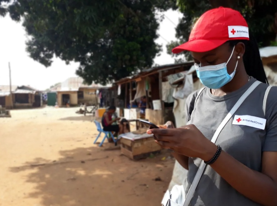 A woman standing outside and wearing a red British Red Cross ball cap and branded t-shirt looks at her phone.